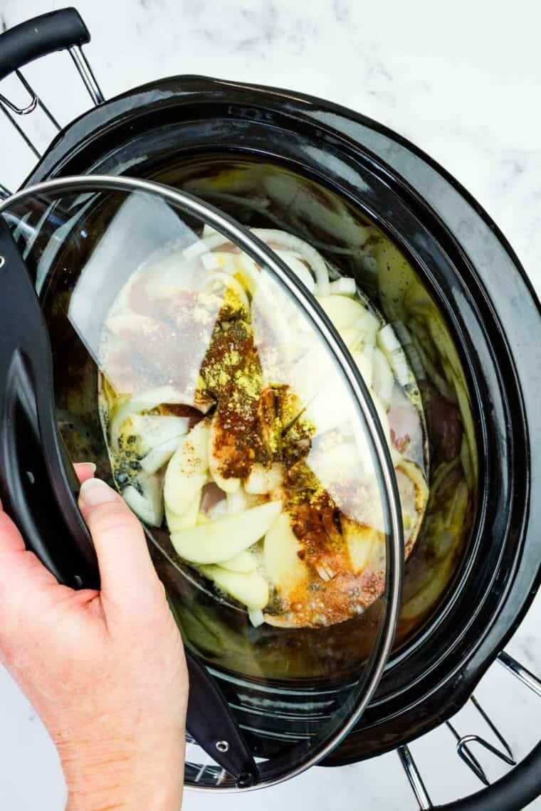 This easy slow cooker pulled chicken is simmered for hours with hard apple cider, apples, and onion. It's SO tender and ideal for sandwiches! Putting the lid on a slow cooker loaded with hard apple cider, chicken, apple slices, onion, and seasonings.