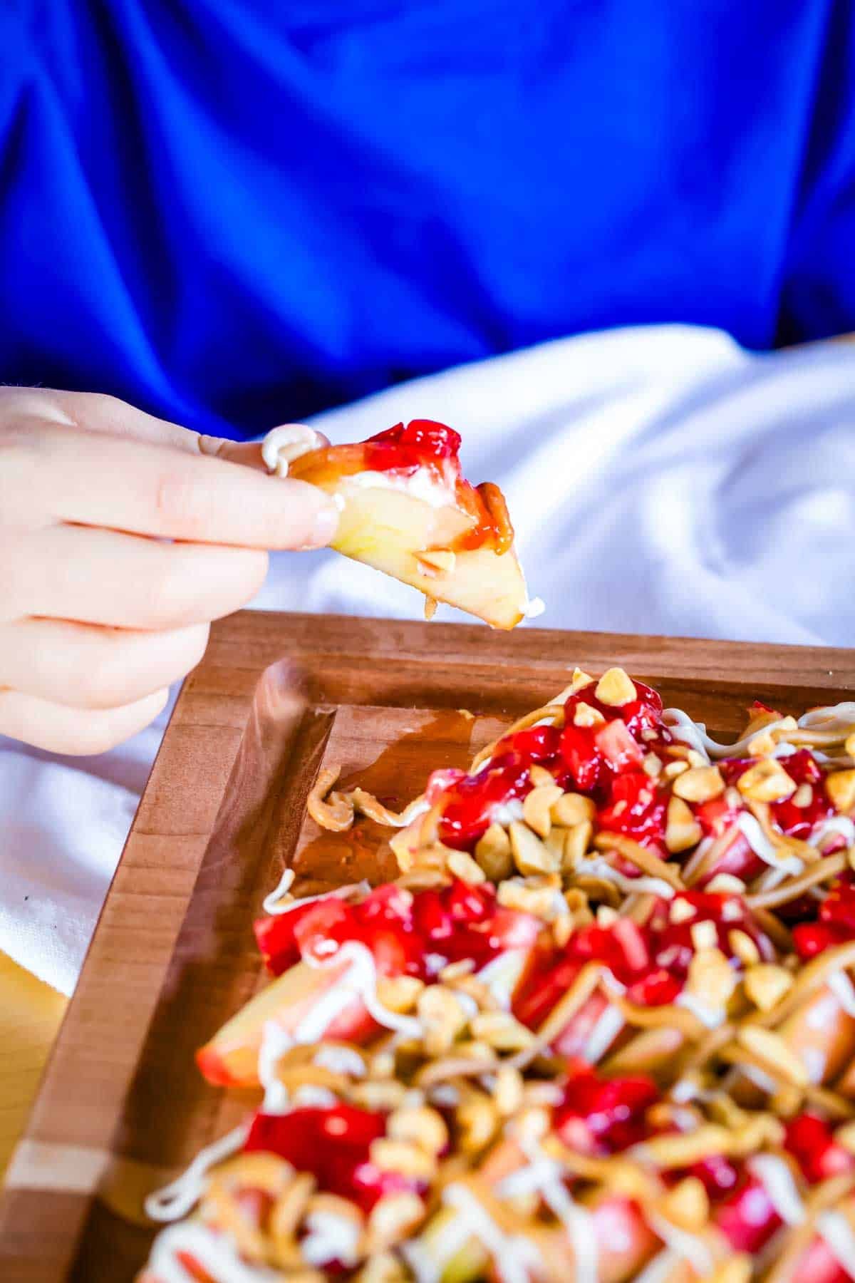 With apple slices instead of chips, no-bake apple nachos are topped with sweetened cream cheese, saucy strawberries, and peanut butter. Yum! A child's hand taking from a platter of apple nachos.