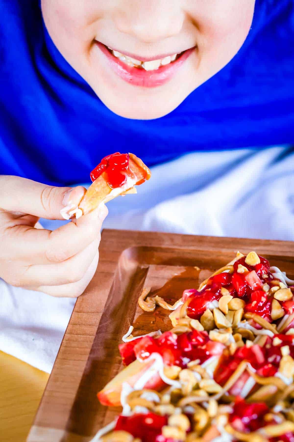 With apple slices instead of chips, no-bake apple nachos are topped with sweetened cream cheese, saucy strawberries, and peanut butter. Yum! A child smiling, eating apple nachos.