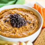 Pumpkin pie hummus in a bowl on a plate surrounded by carrots, apples, and crackers.