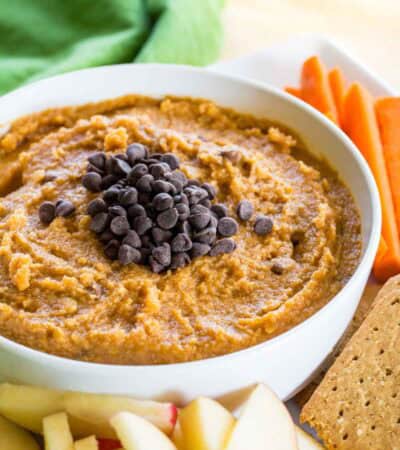 Pumpkin pie hummus in a bowl on a plate surrounded by carrots, apples, and crackers.