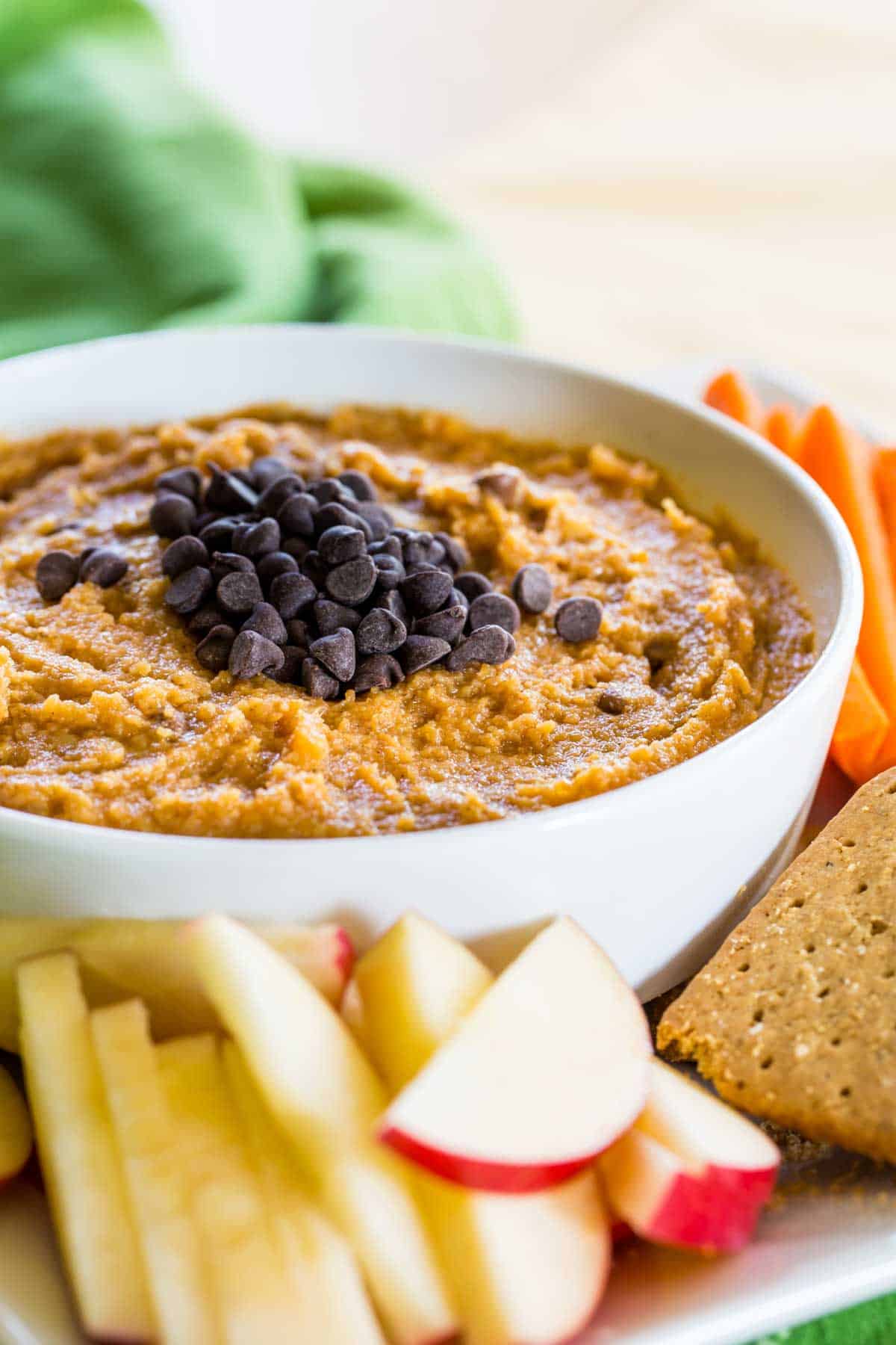 Pumpkin hummus in a bowl on a plate loaded with dippables (carrots, apples, crackers).
