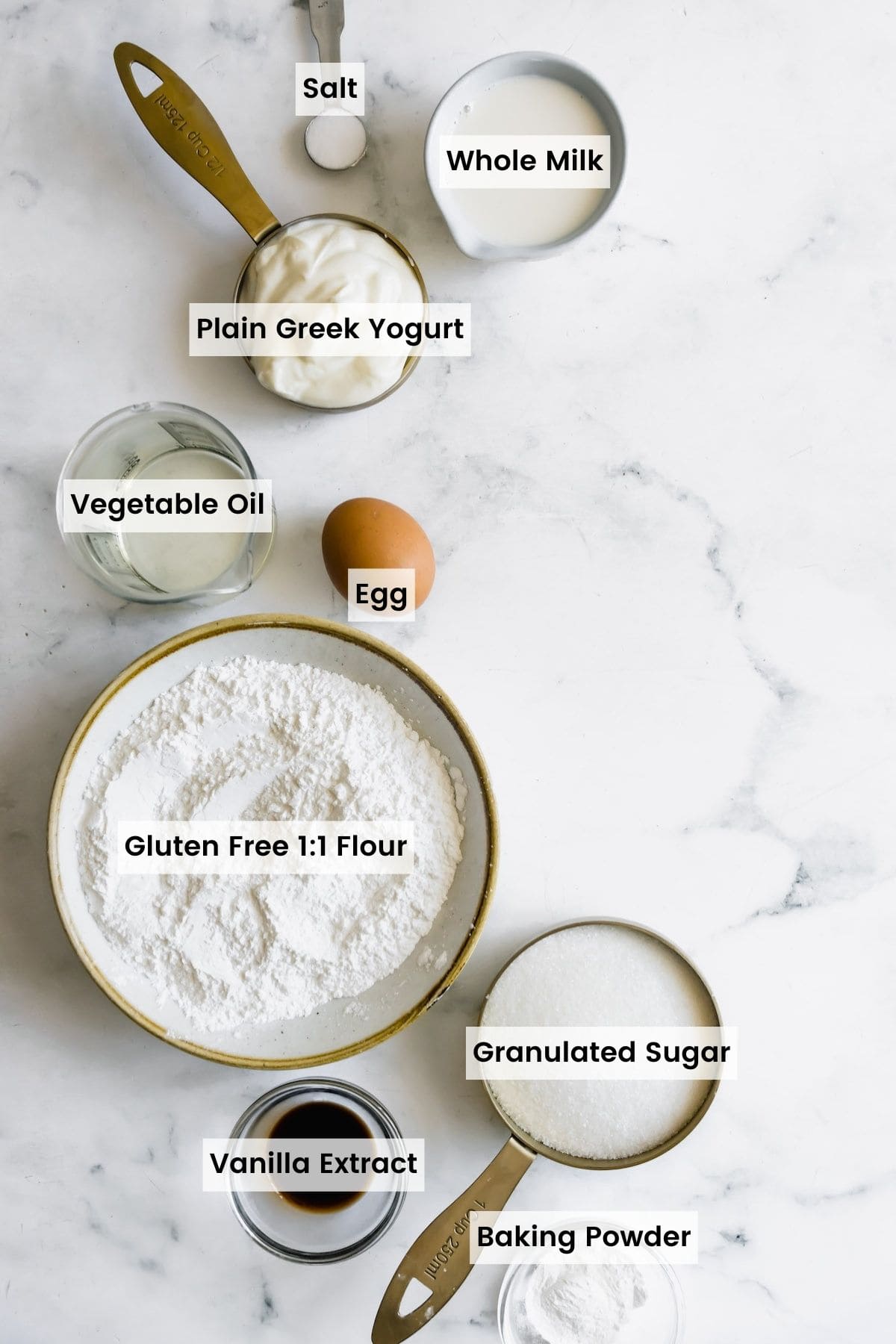Flour, An Egg, Vanilla Extract and the Other Cupcake Ingredients Arranged on a Marble Countertop with text labels.