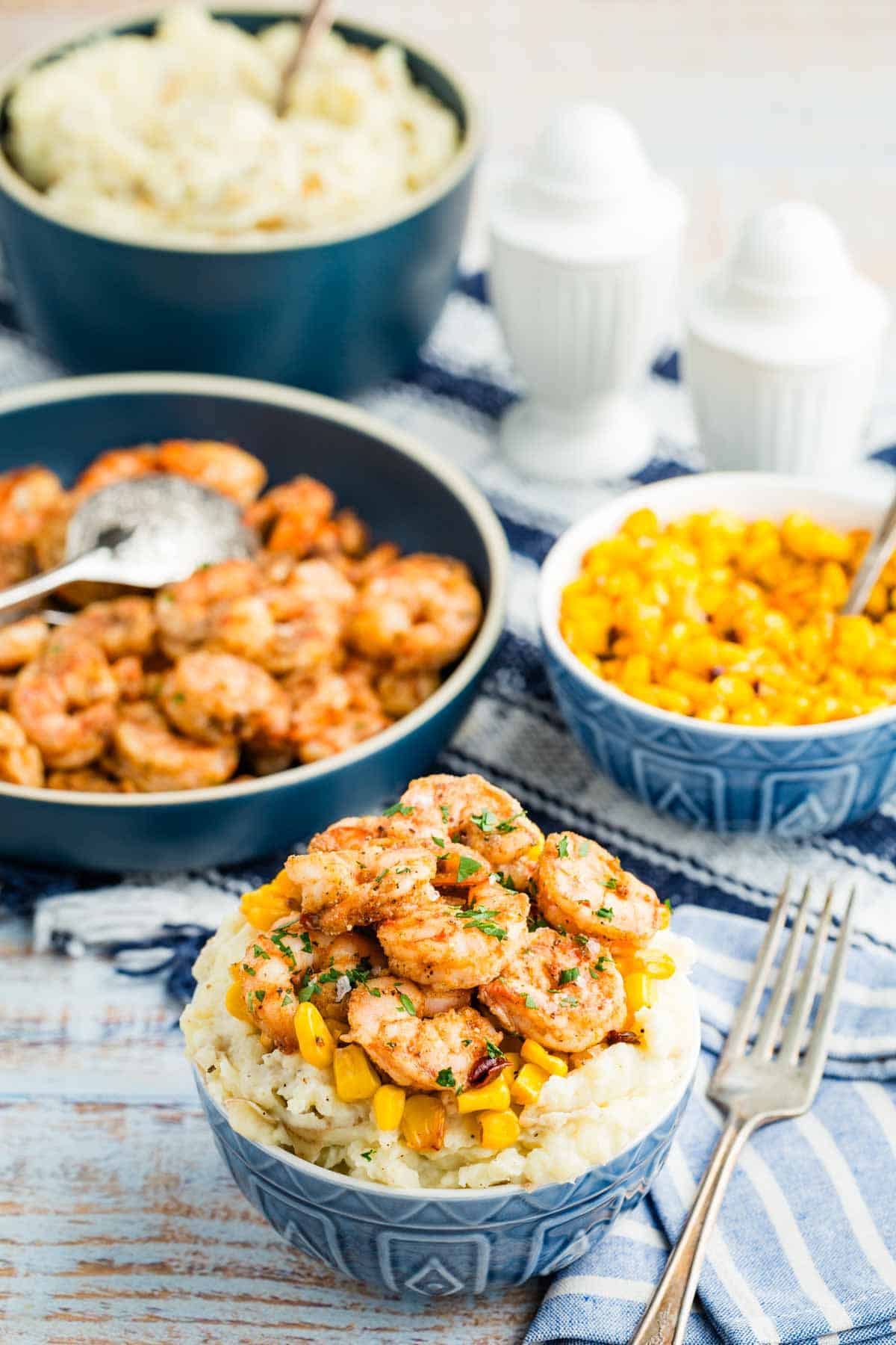 Shrimp and mashed potatoes in a bowl with additional shrimp, potatoes, and corn in the background.