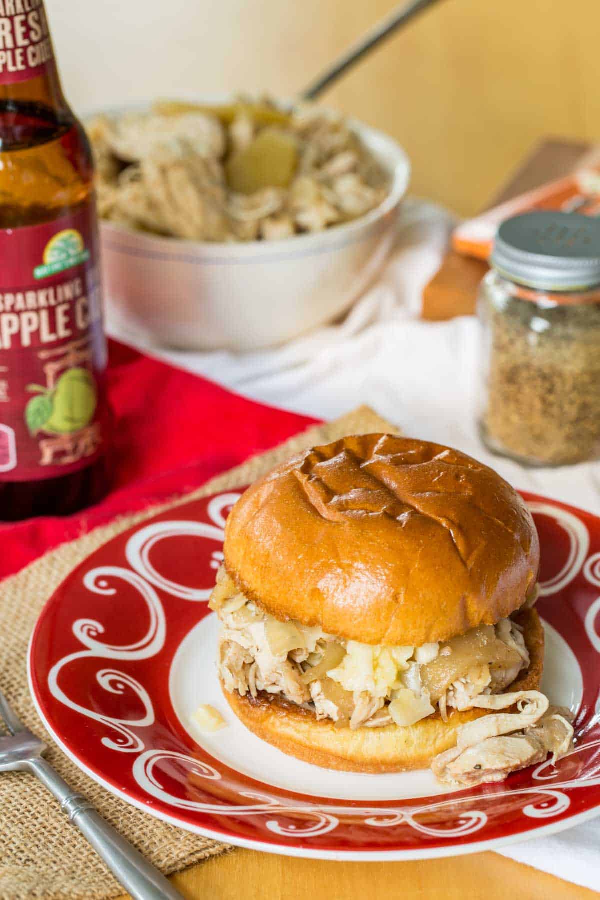 This easy slow cooker pulled chicken is simmered for hours with hard apple cider, apples, and onion. It's SO tender and ideal for sandwiches! Shredded chicken, apples, and cheese on a bun on a red and white plate.
