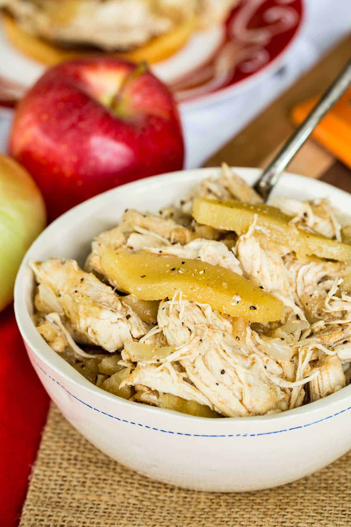 This easy slow cooker pulled chicken is simmered for hours with hard apple cider, apples, and onion. It's SO tender and ideal for sandwiches! A bowl of shredded chicken with apple slices.