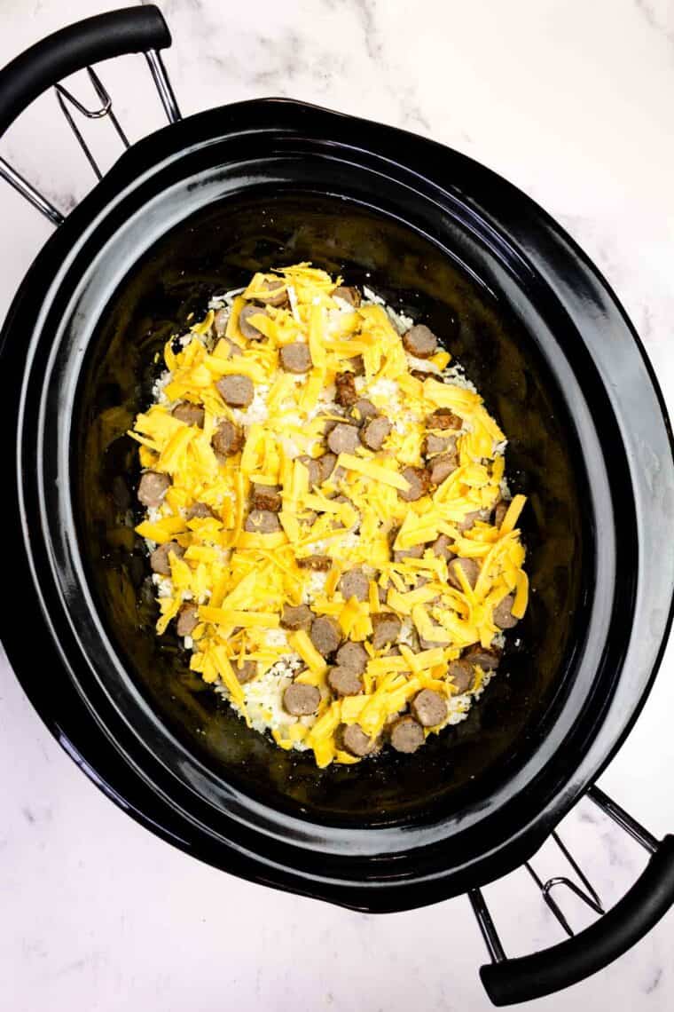 Adding layers of shredded cauliflower, sausage, and cheddar cheese to a slow cooker.