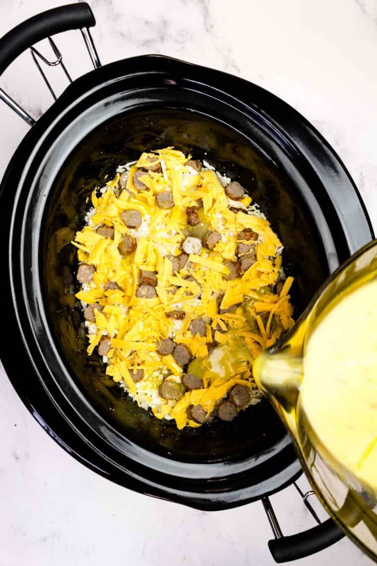 Pouring eggs over layers of shredded cauliflower, sausage, and cheddar cheese in a slow cooker.