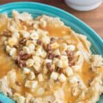 Apple hummus spread in a turquoise oval dish topped with caramel, apple pieces, chopped pecans, and white chocolate chips.