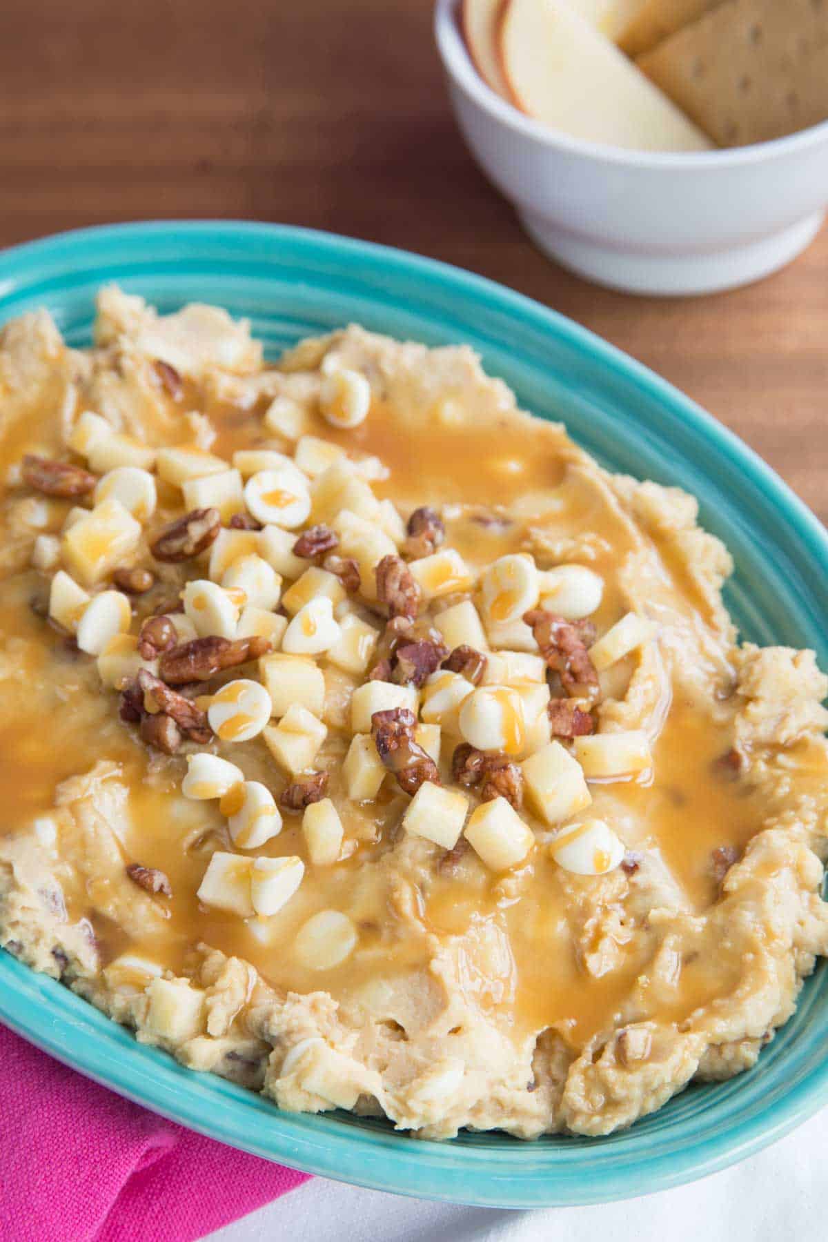 Apple hummus spread in a turquoise oval dish topped with caramel, apple pieces, chopped pecans, and white chocolate chips.