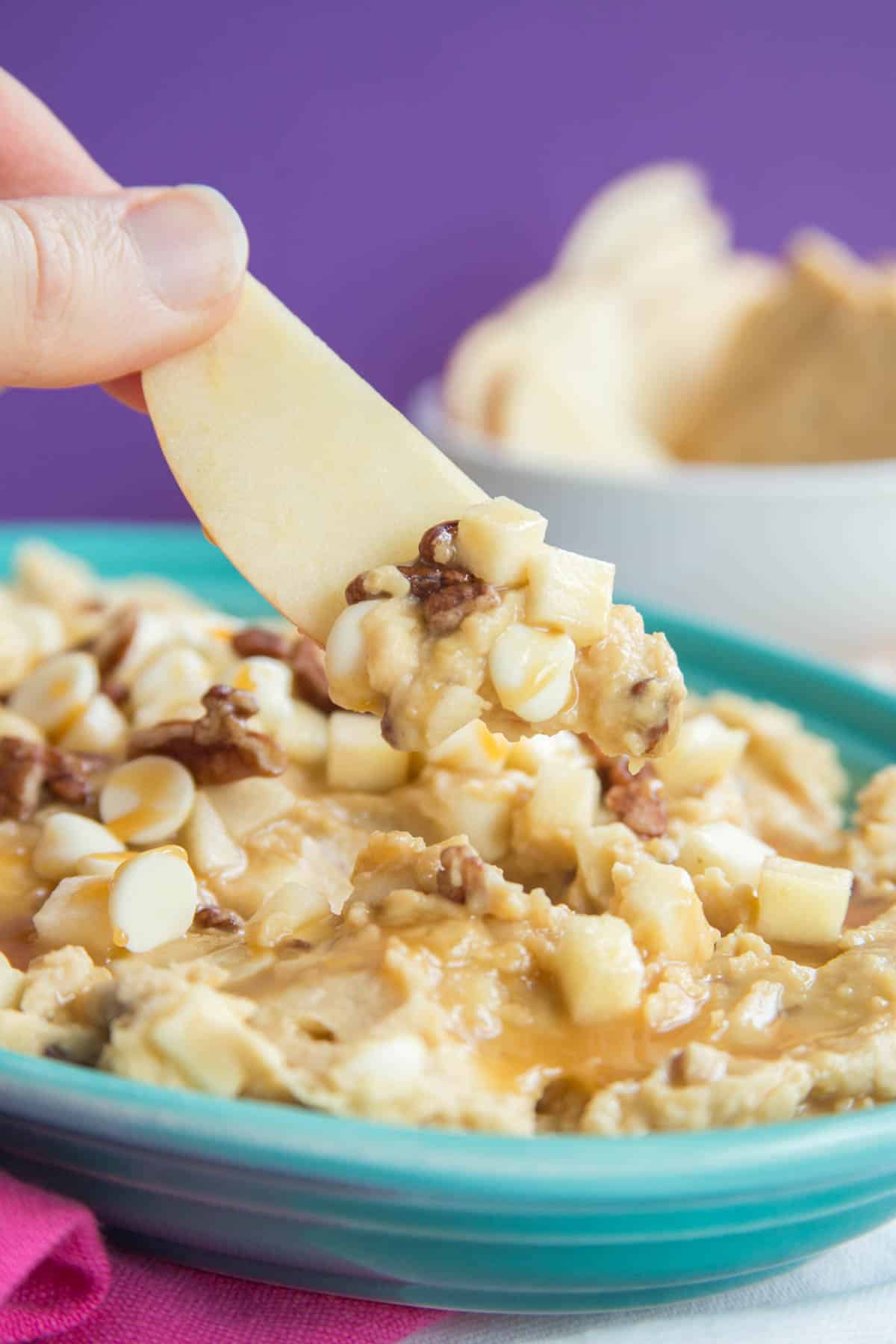 Apple hummus topped with caramel, pecans, white chocolate chips, and apple pieces scooped up on an apple slice.