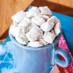 White chocolate muddy buddies snack mix in a blue mug with text overlay that says "Chai White Chocolate Puppy Chow".