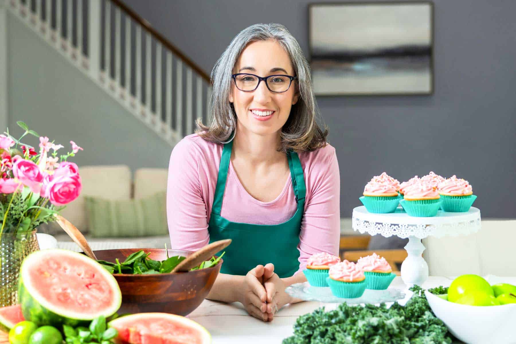 A woman in glasses leaning on a countertop with cupcakes, greens, and other food items around her.