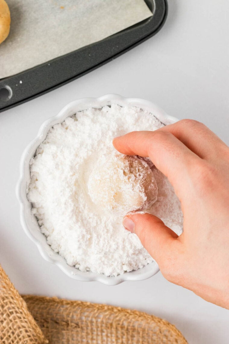 Rolling a gluten-free white chocolate macadamia snowball cookie in powdered sugar.