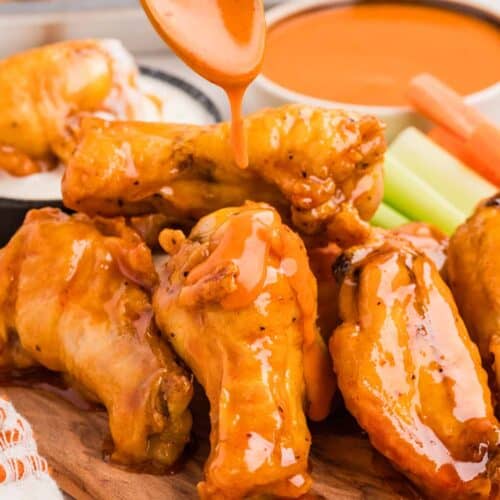 Drizzling buffalo sauce over baked buffalo wings on a wooden baking sheet