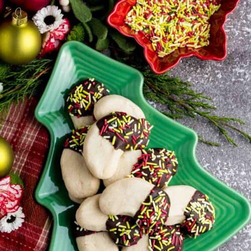 An overhead view of a Christmas tree-shaped dish with almond crescent cookies on it next to a dish of sprinkles.