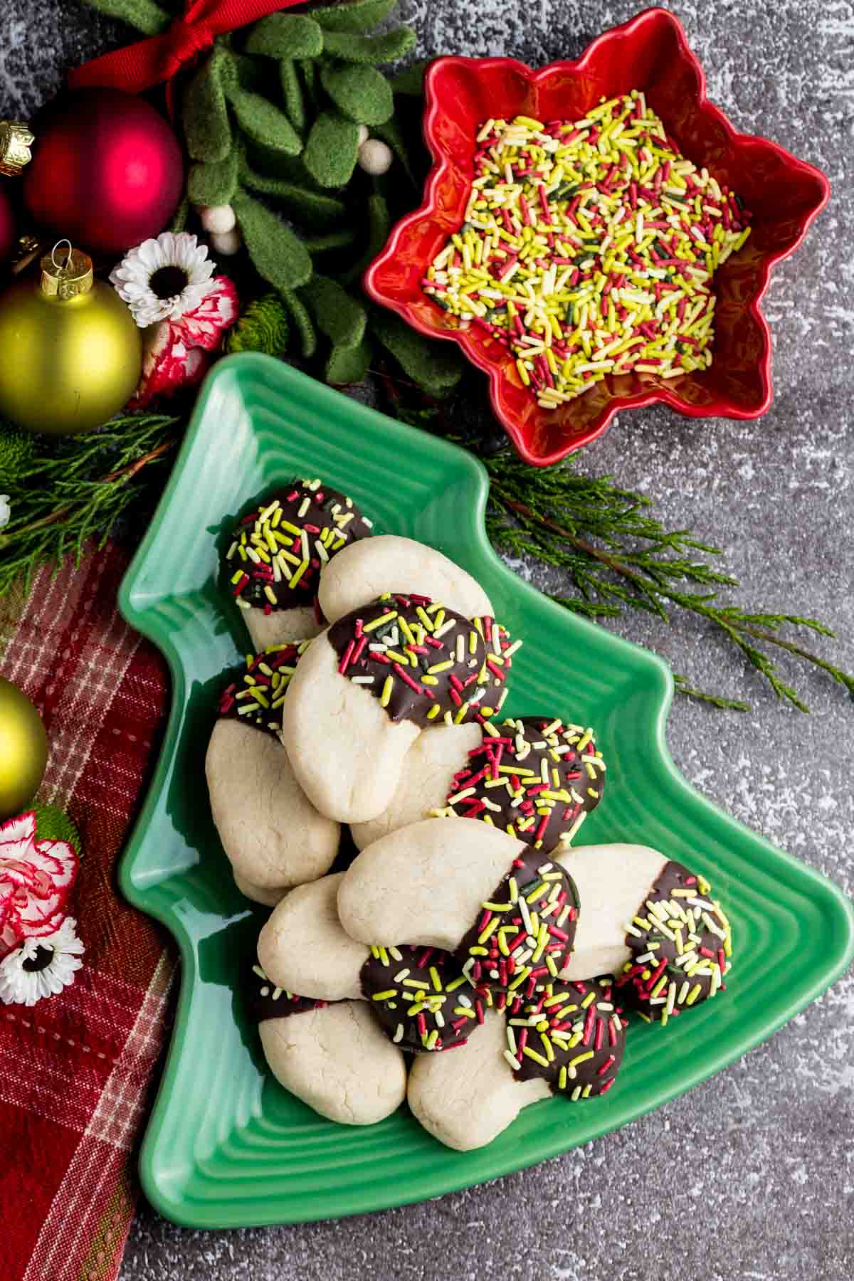 An overhead view of a Christmas tree-shaped dish with almond crescent cookies on it next to a dish of sprinkles.
