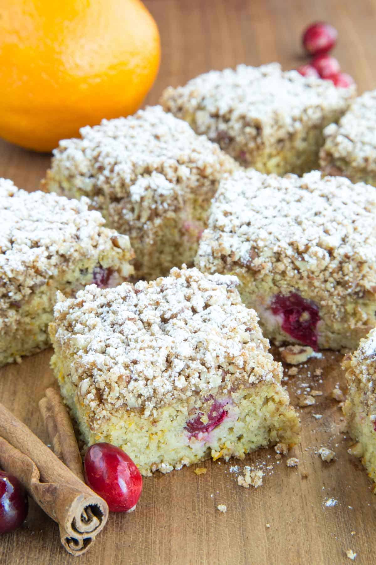 Pieces of gluten free cranberry cake on a cutting board with an orange, cinnamon sticks, and fresh cranberries around them.