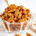 A glass bowl of candied walnuts with a spoon in it and some spilled out in front.