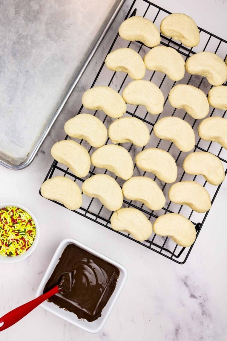 Baked almond crescent cookies on a rack next to bowls of melted chocolate and sprinkles.