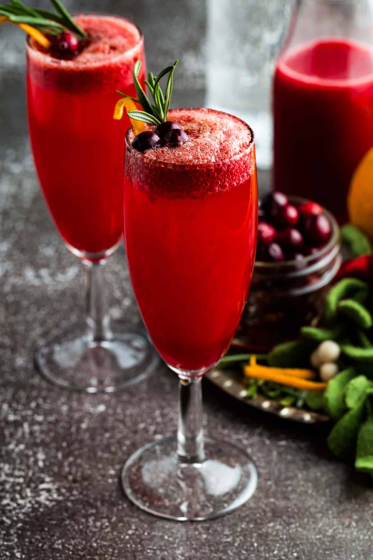 Two flute glasses with a cranberry mocktail garnished with cranberries, orange zest, and rosemary.