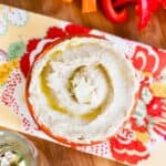 A birds eye view of a bowl of white bean hummus on a cutting board with veggies.