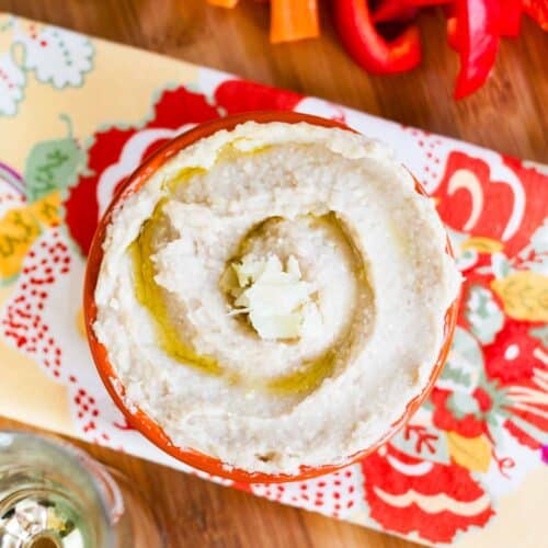 A birds eye view of a bowl of white bean hummus on a cutting board with veggies.