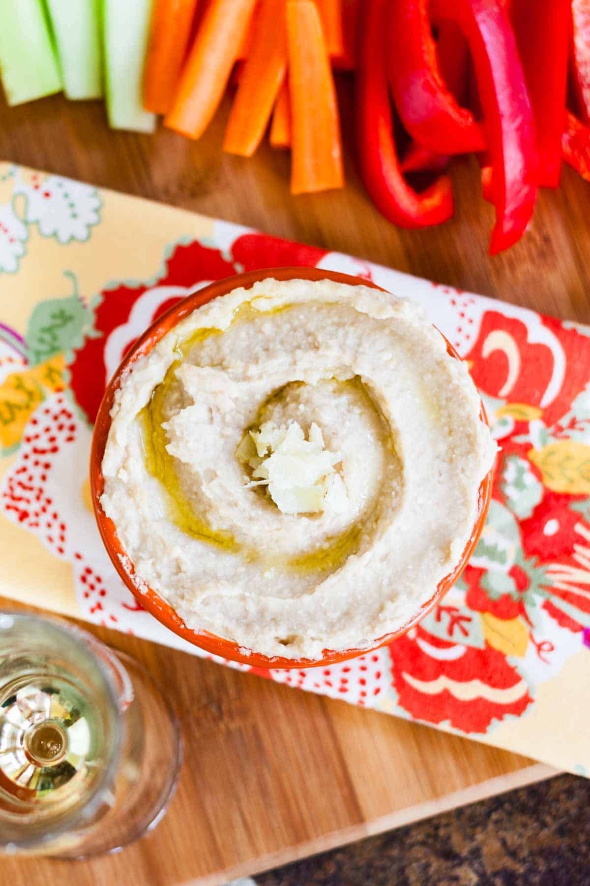 A birds eye view of a bowl of white bean hummus on a cutting board with veggies.
