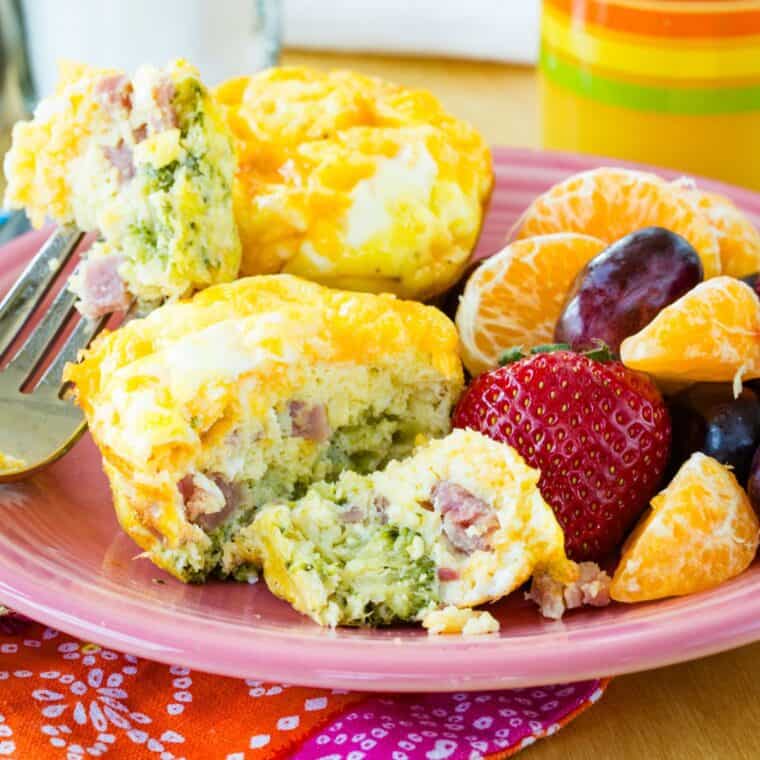 Broccoli ham and cheese egg muffins on a plate with one cut into with a fork and fruit on the plate.