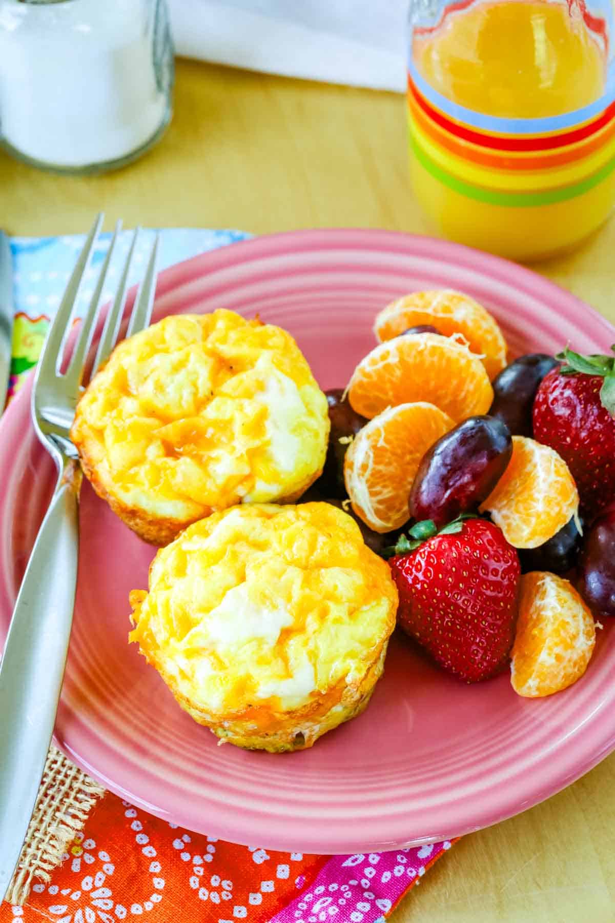 An overhead view of two egg muffins, fruit, and a fork on a pink plate.