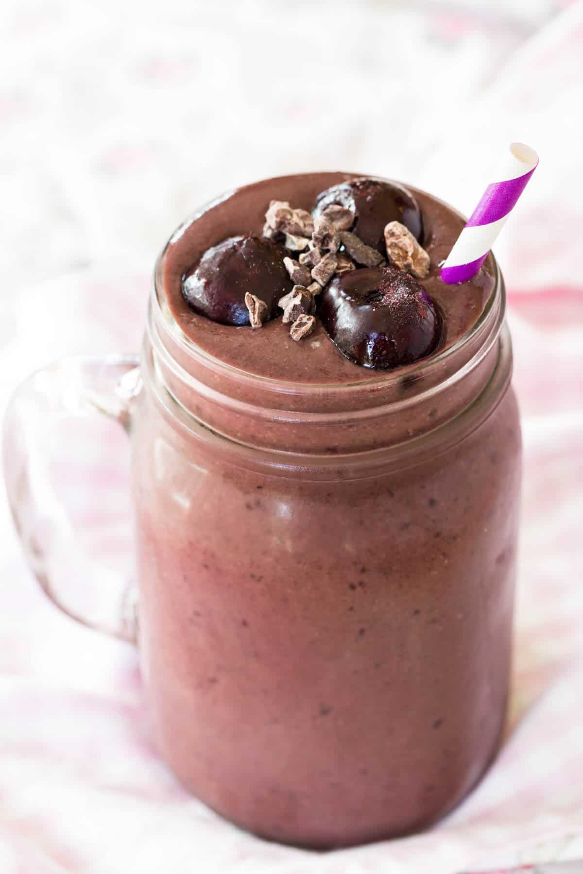 A chocolate cherry smoothie in a glass jar with a striped straw and frozen cherries on top.