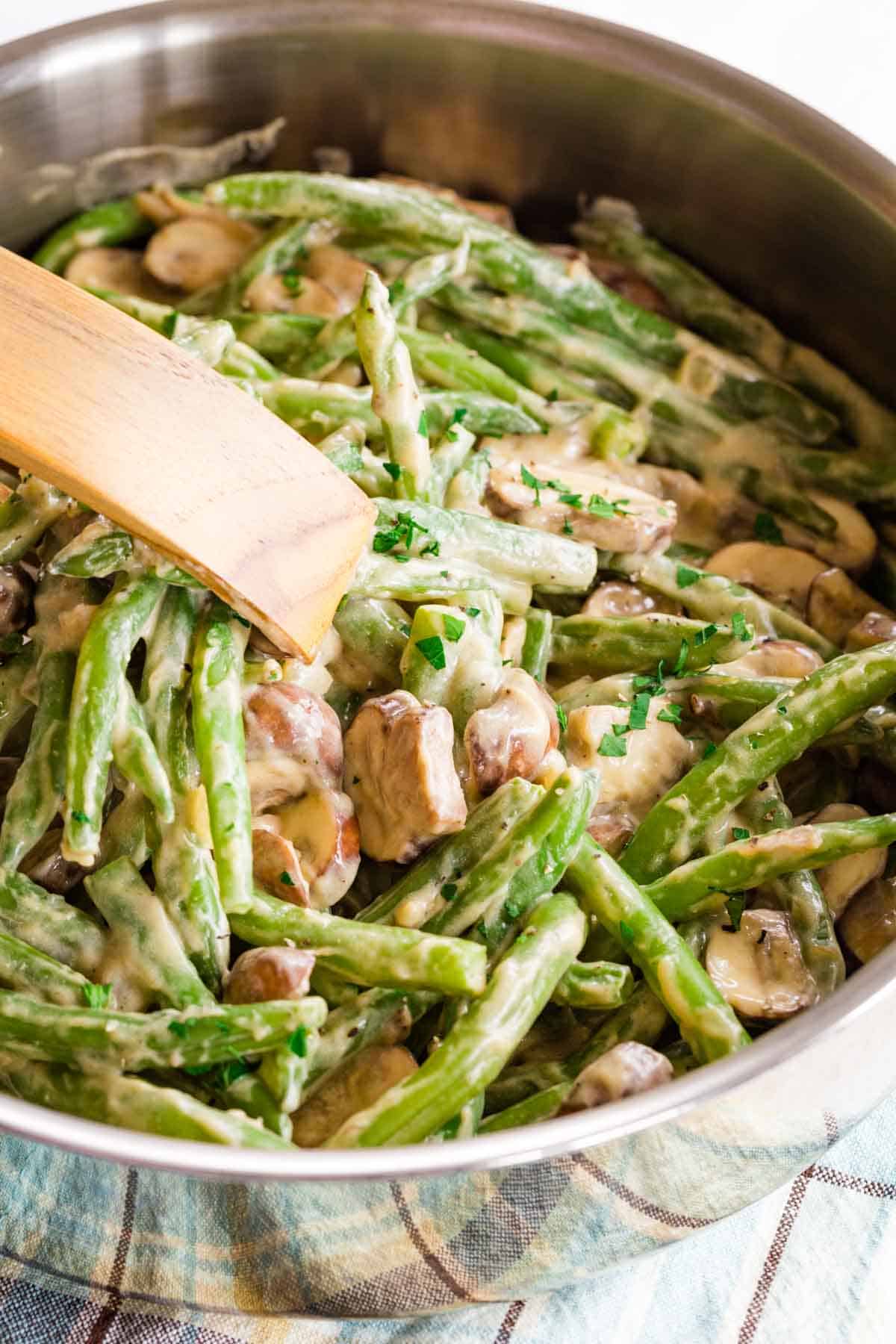 A close-up of green beans and mushrooms with a creamy sauce being picked up with a wooden pair of tongs from a skillet.