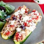 Two zucchini halves filled with chicken, tomatoes, olives, and feta on a plate with text overlay that says "Greek Chicken Stuffed Zucchini".