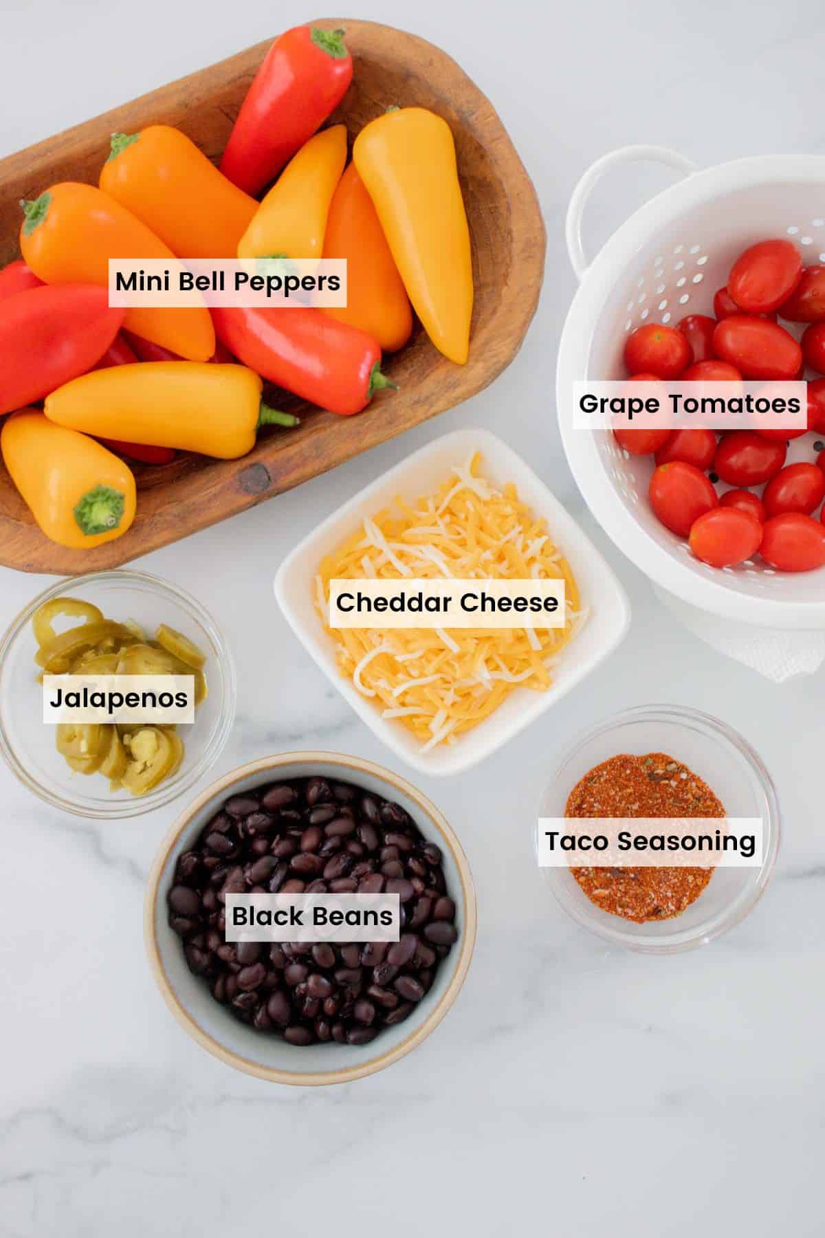 Bowls of mini bell peppers, taco seasoning, black beans, grape tomatoes, jalapeno slices, and shredded cheese with text labels.