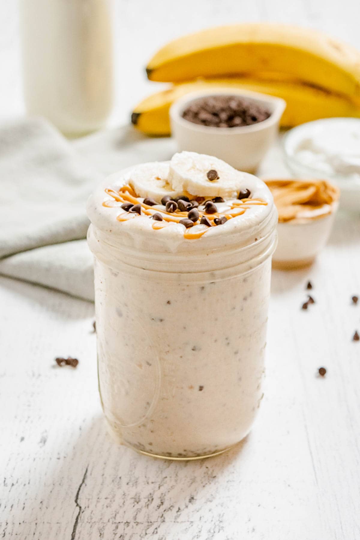 A peanut butter smoothie in a glass jar with bananas and bowld of peanut butter and mini chocolate chips in the background.