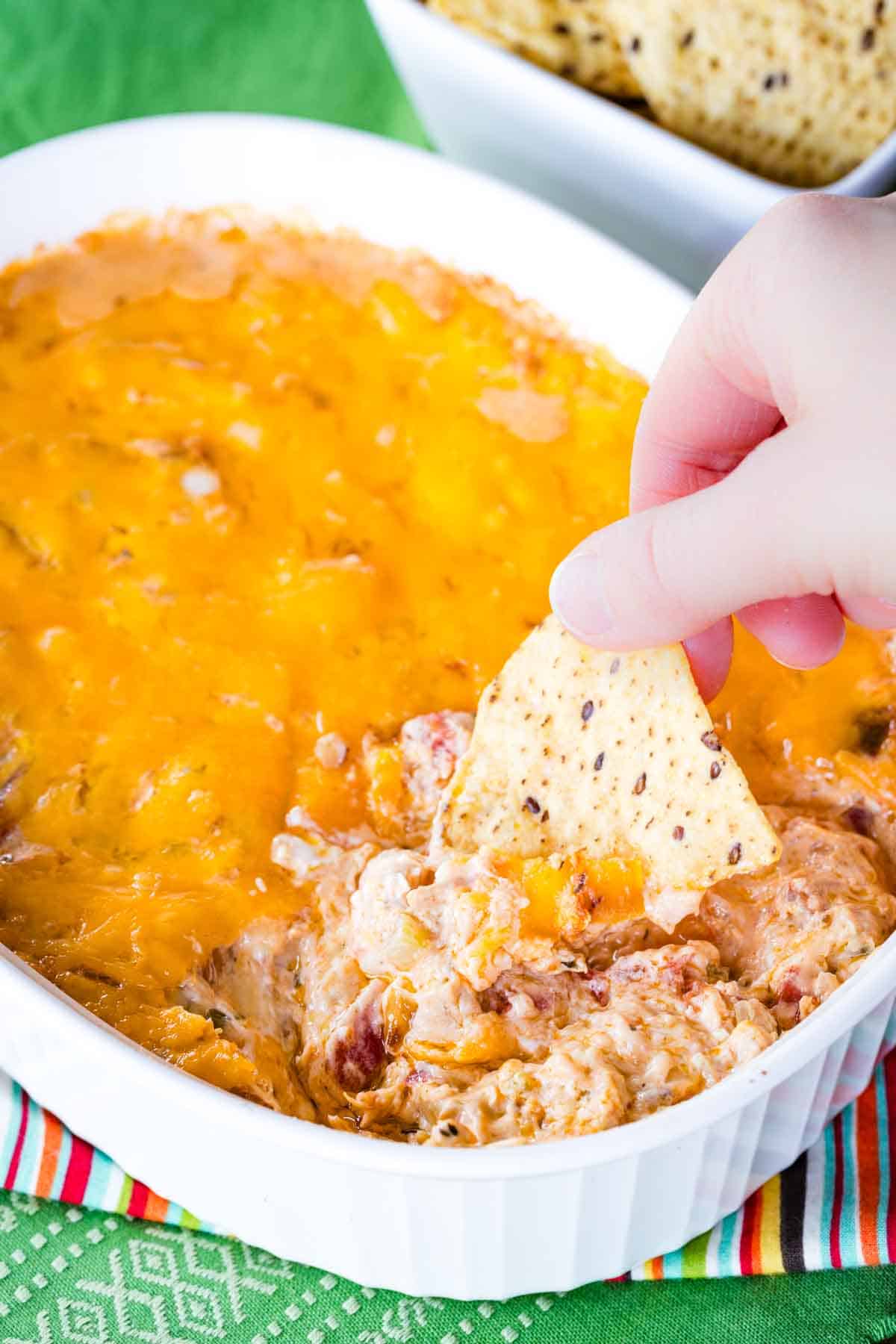 Low Carb Jambalaya Dip - a hot and cheesy party appetizer recipe with chicken, shrimp, Andouille sausage and Cajun seasoning plus plenty of cheese! #cheesy #dip #snack #keto Cheese topped jambalaya dip in a baking dish with a hand reaching in holding a tortilla chip.