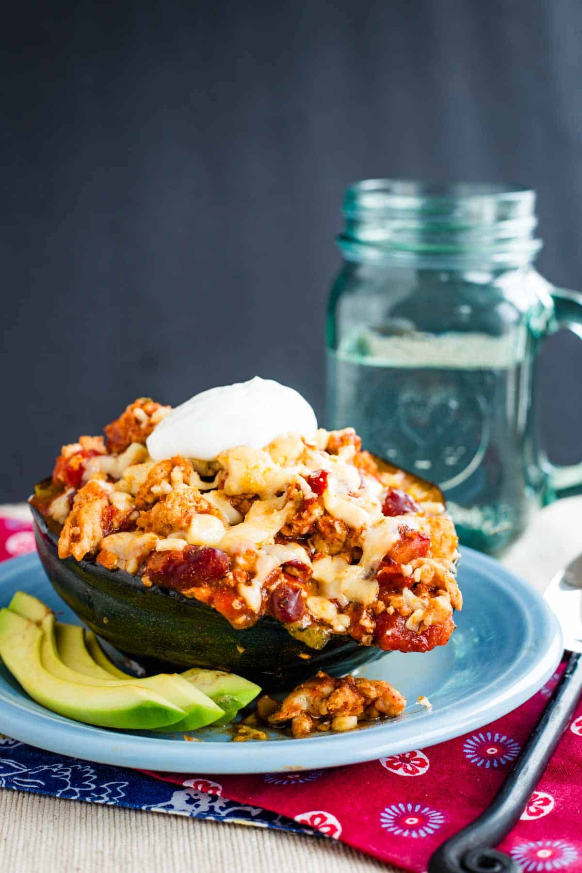 Chili overflowing out of a half of a roasted acorn squash in on a plate next to some avocado slices.