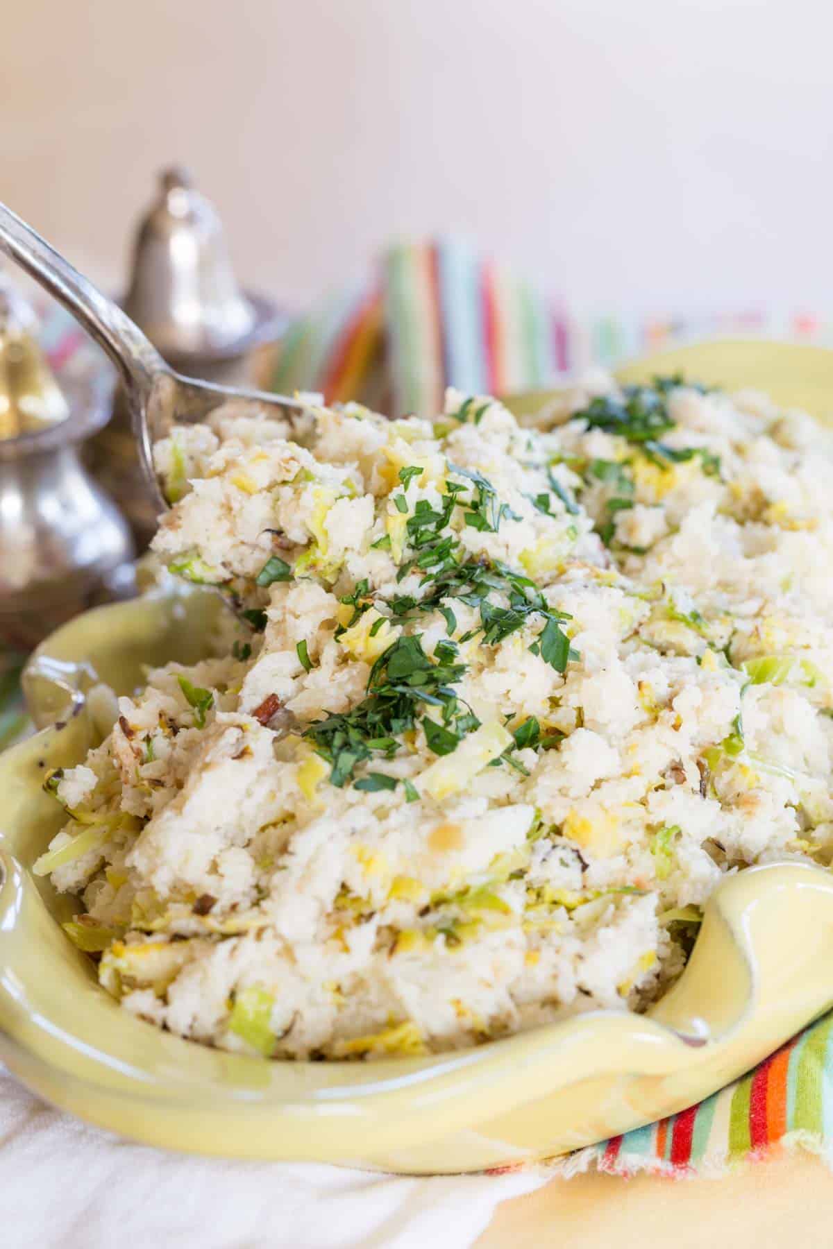 Spoon scooping cauliflower colcannon out of a yellow serving dish.