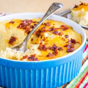 Cheesy mashed cauliflower bake topped with crumbled bacon in a casserole dish with a serving spoon in it.