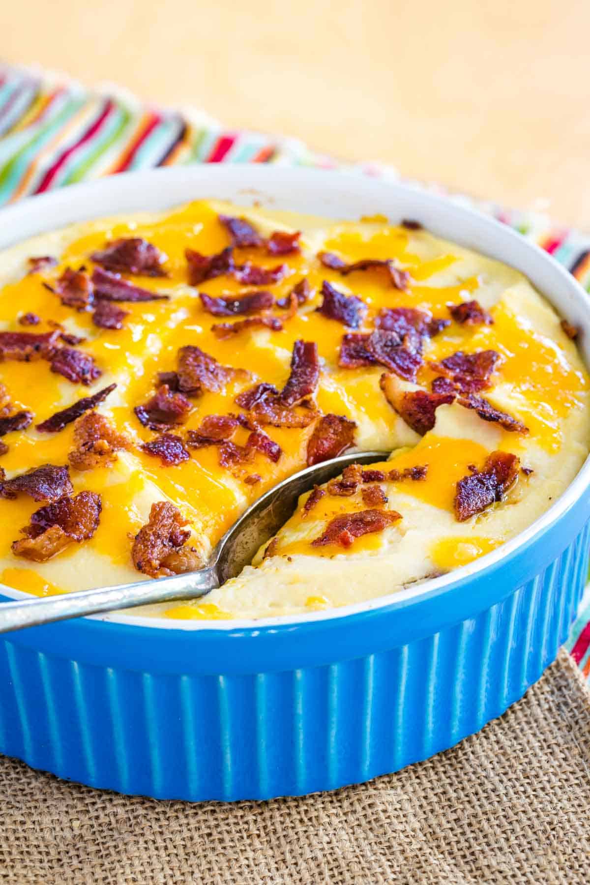 A spoon resting in a round blue baking dish filled with creamy cauliflower mash topped with bacon and cheese.