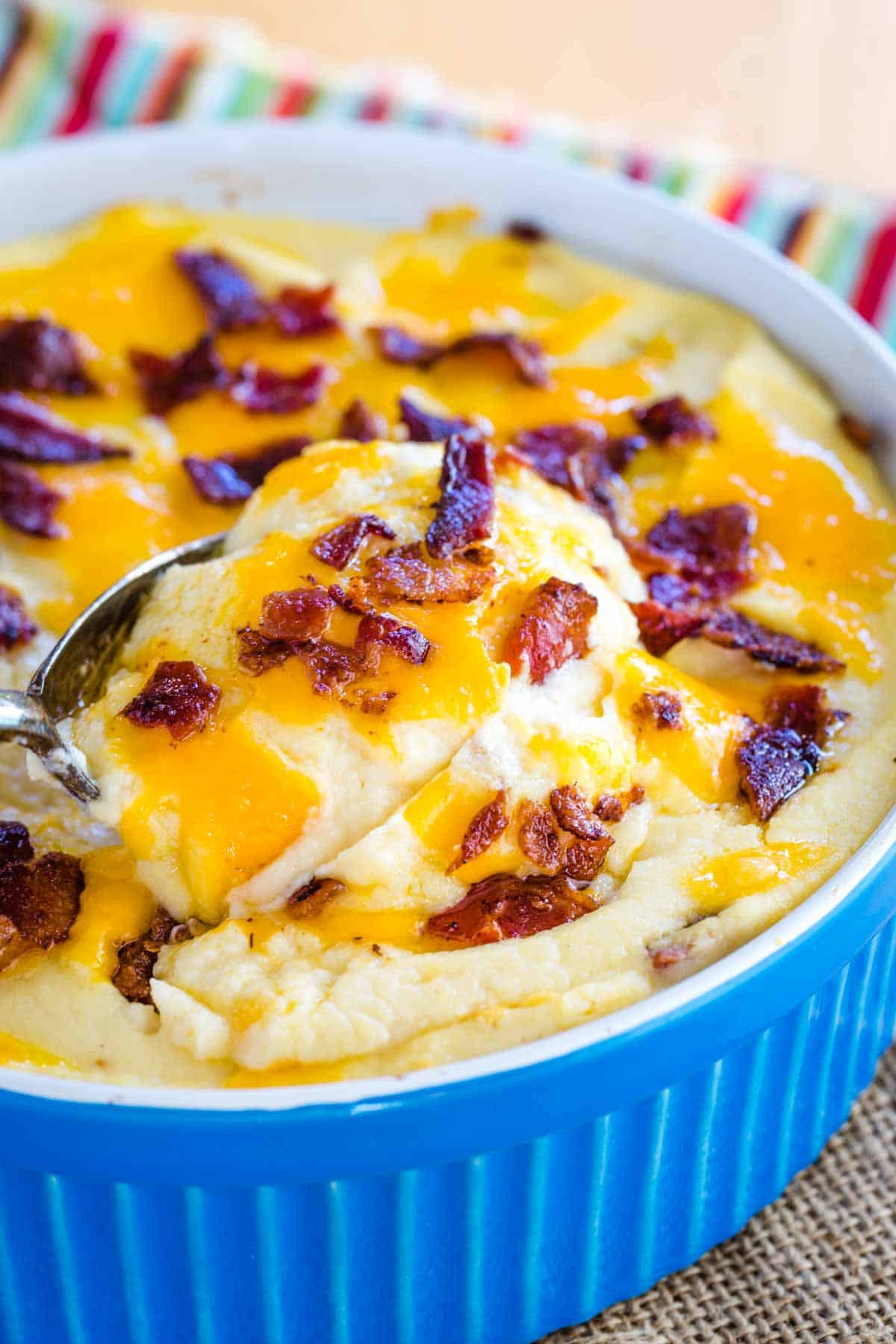 A spoon scooping up creamy mashed cauliflower topped with bacon and cheese from a blue dish.