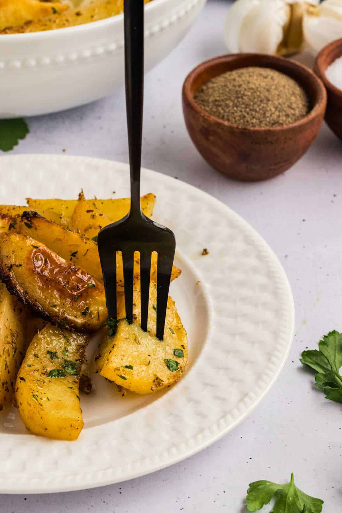 Potatoes are baked in a lemony, garlicky broth until golden on the outside and soft on the inside in this Greek Potatoes recipe. A fork in a Greek potato on a white plate
