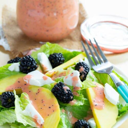 A mango blackberry goat cheese salad on a plate with a fork with a jar of strawberry poppyseed dressing behind it.