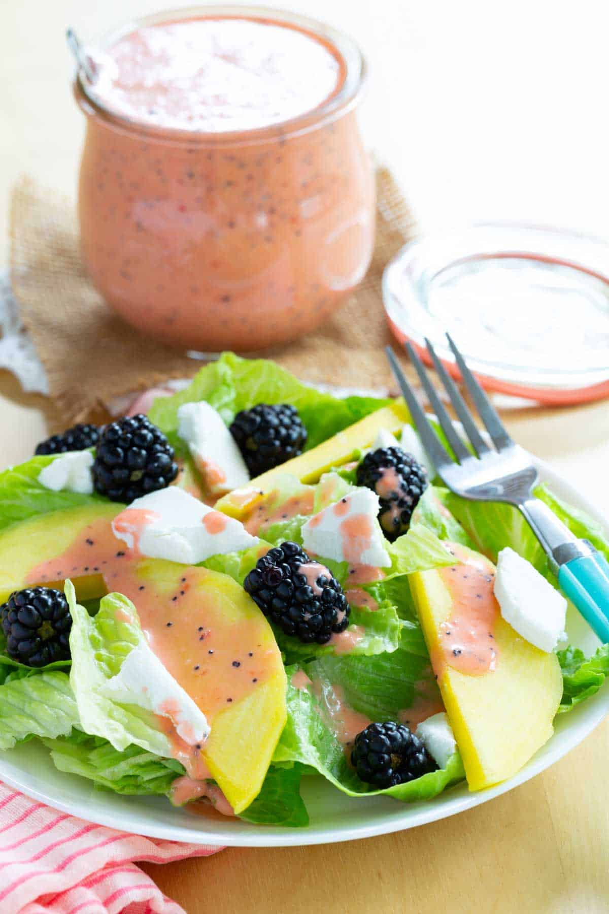 A mango blackberry goat cheese salad on a plate with a fork with a jar of strawberry poppyseed dressing behind it.