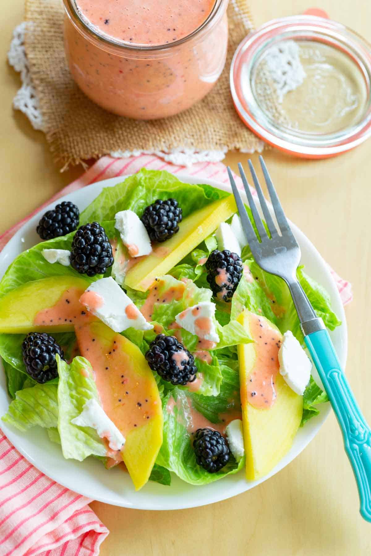 A birds eye view of a plate of lettuce topped with mango slices, blackberries, goat cheese, and a pink poppyseed dressing.