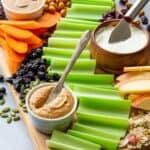Celery, apples, carrots, bowls of peanut butter and cream cheese, and other toppings on a wooden board.