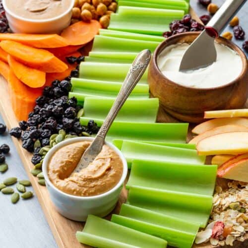 Celery, apples, carrots, bowls of peanut butter and cream cheese, and other toppings on a wooden board.