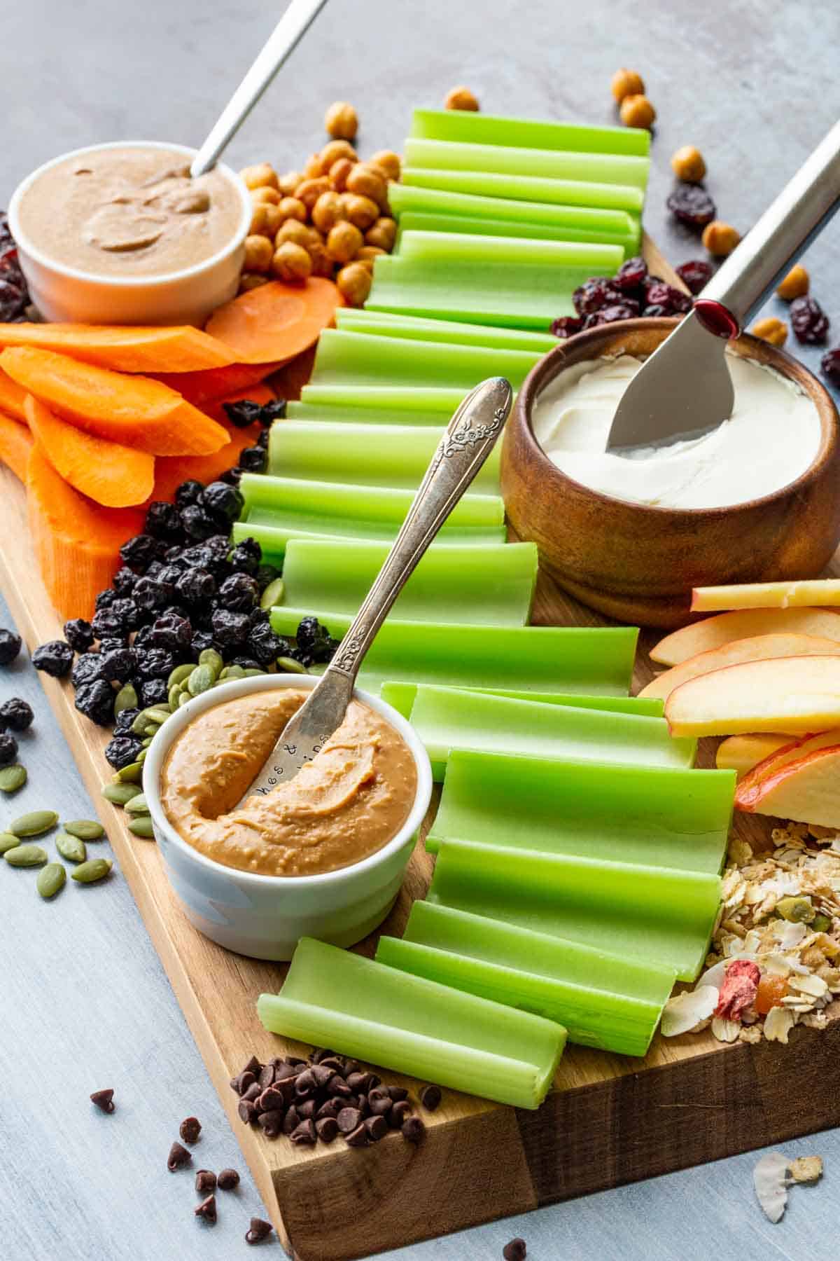 Celery, apples, carrots, bowls of peanut butter and cream cheese, and other toppings on a wooden board.