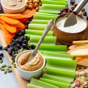 A cutting board filled with celery stalks, a bowl of peanut butter, and other fruits, veggies, and toppings to make ants on a log.
