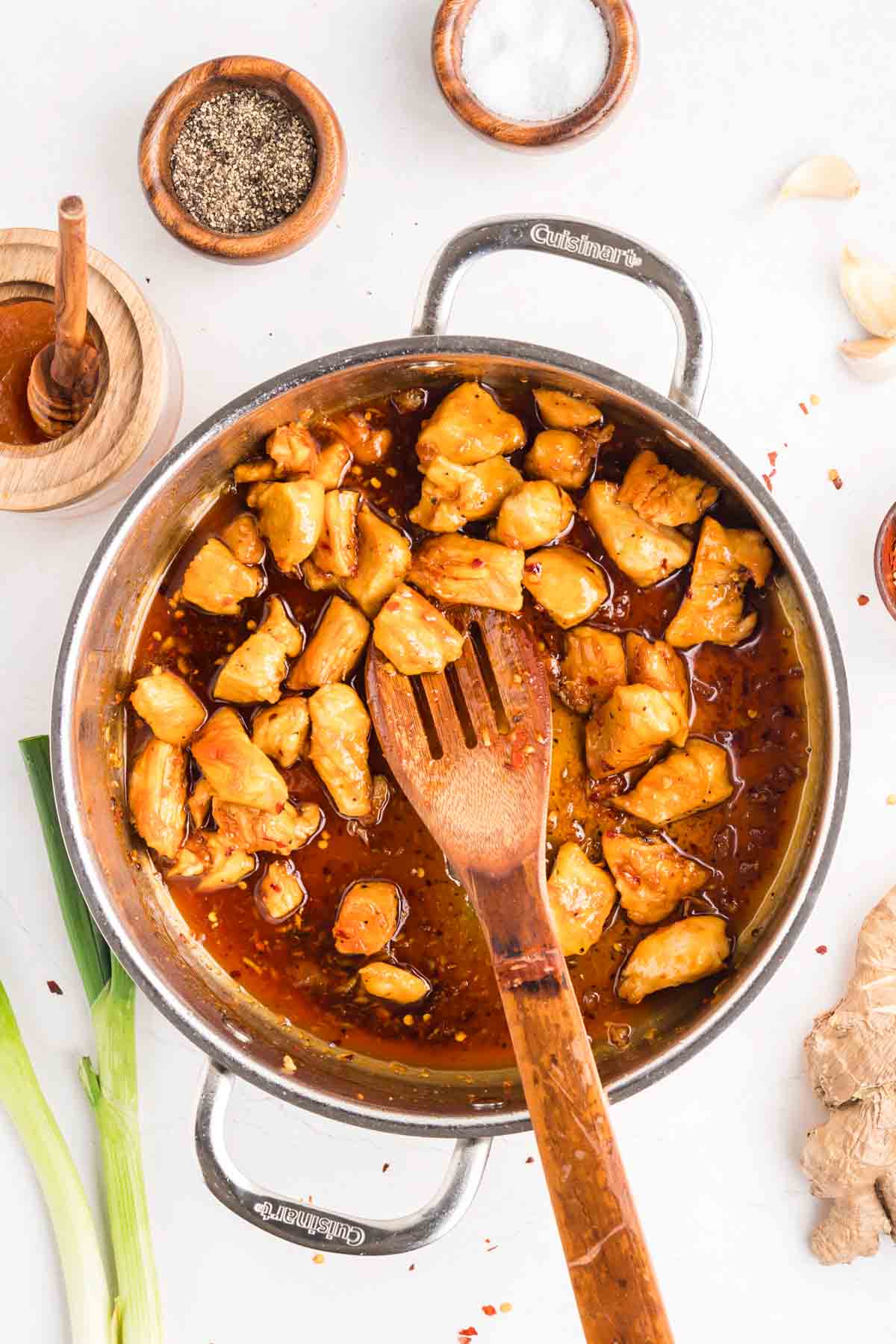 This easy sweet and spicy chicken is full of flavor, with honey, brown sugar, and sriracha. Ready in just 20 minutes! A wooden spoon stirring sweet and spicy chicken in a pan